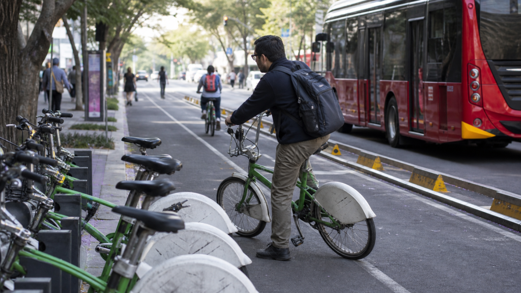 Persona usando una bicicleta pública ECOBICI junto a una cicloestación y una avenida de Ciudad de México.